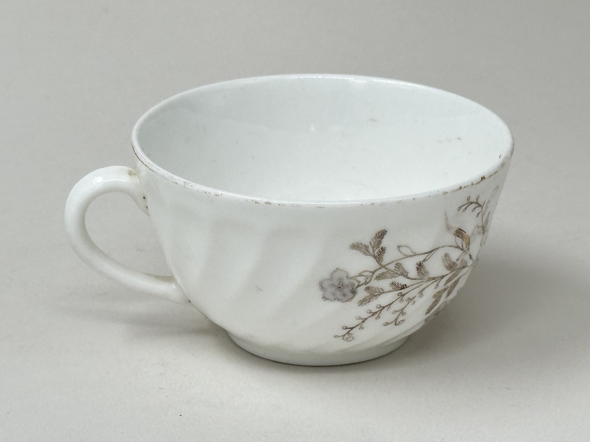 Koppar i vitt fältspatsporslin med tryckt blomdekor i brunt, guld och grått. Guldrand längs mynning och öra (delvis avskavt). Vriden godronnering runtom.