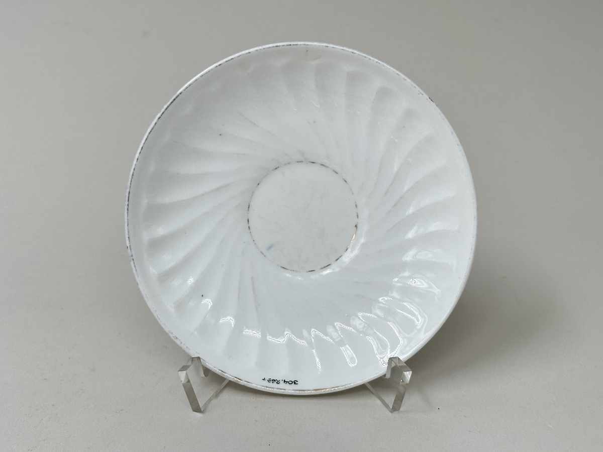 Kaffefat i vitt fältspatsporslin, med guldrand längs ytterkant och koppytans kant (delvis avskavt). Vriden godronnering runt brätte.