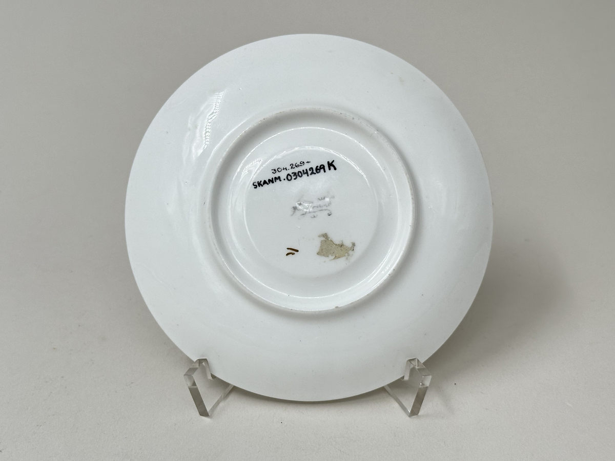 Kaffefat i vitt fältspatsporslin, med guldrand längs ytterkant och koppytans kant (delvis avskavt). Vriden godronnering runt brätte.