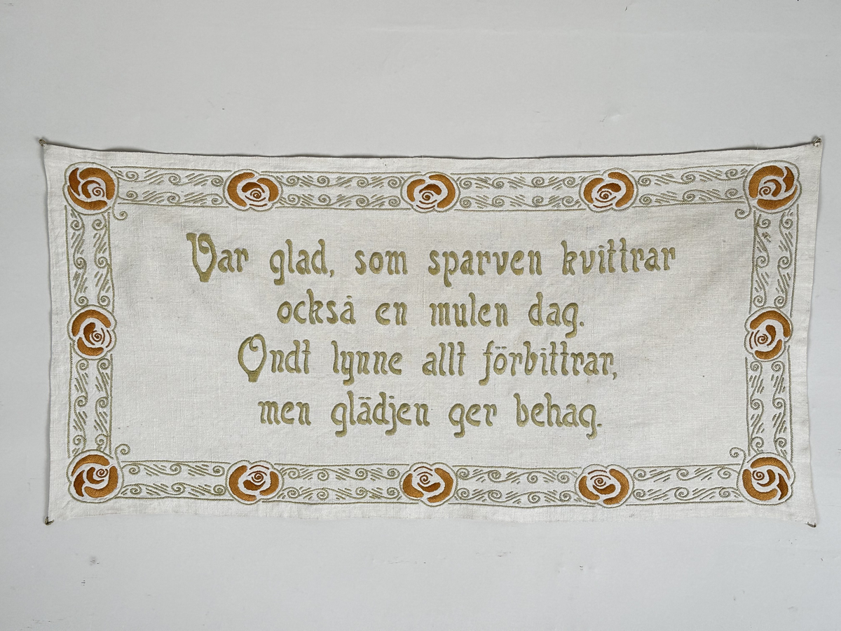 Väggbonad i vit linnelärft. Broderi i pärlgarn, stjälksöm och plattsöm (klumpsöm). Bård runt om med vågmönster i ljusgrönt, blommor i orange och rött. 