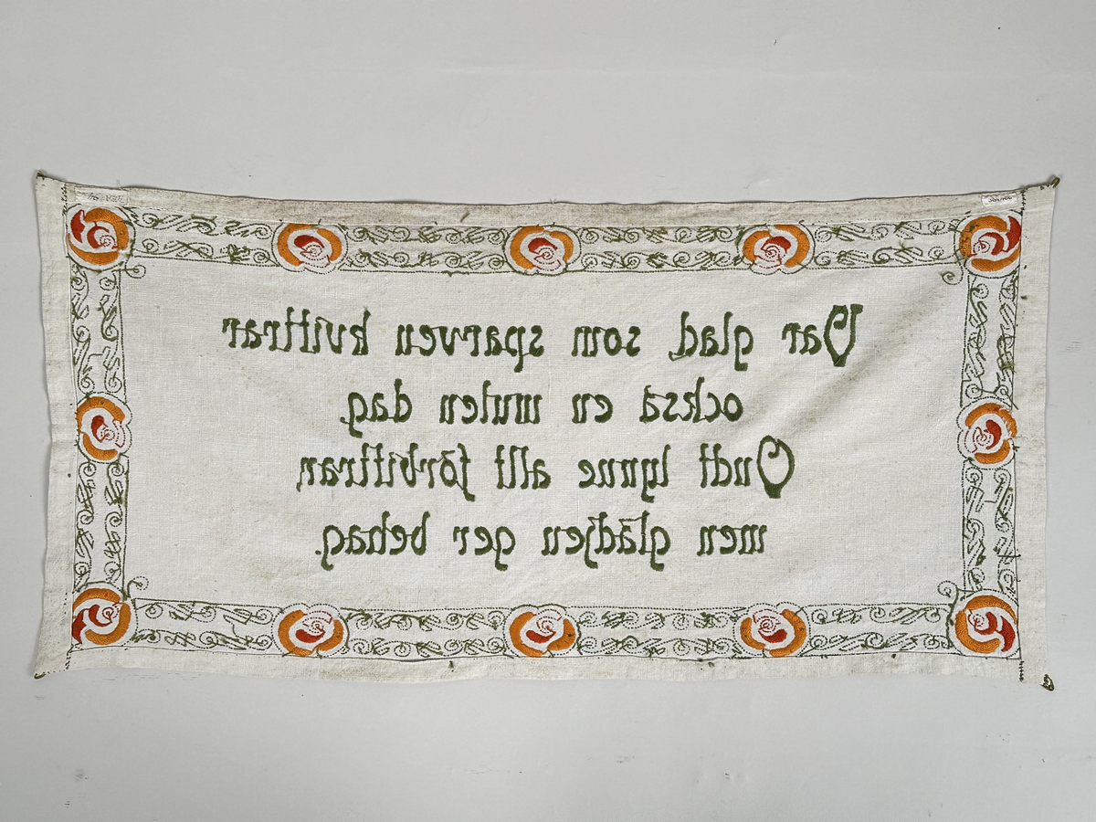 Väggbonad i vit linnelärft. Broderi i pärlgarn, stjälksöm och plattsöm (klumpsöm). Bård runt om med vågmönster i ljusgrönt, blommor i orange och rött. 