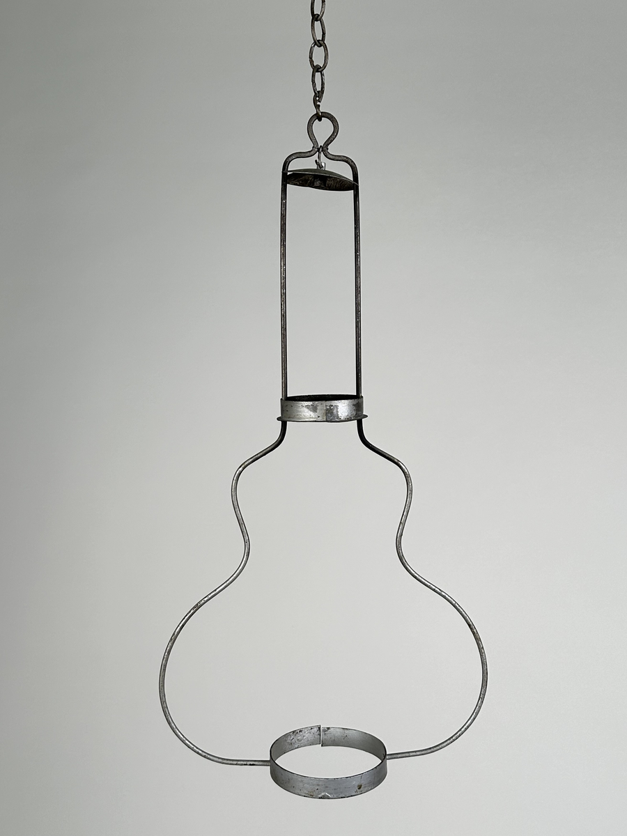 Hängande ställning för fotogenlampa, av två grova metalltrådar som bildar en gitarrform, med ringar av bandplåt nertill samt på halsen för att stödja lampan. Rökhatt med två urtag för metalltrådarna fäst högst upp i ställningen. Silvermålad. Hänger i kätting.