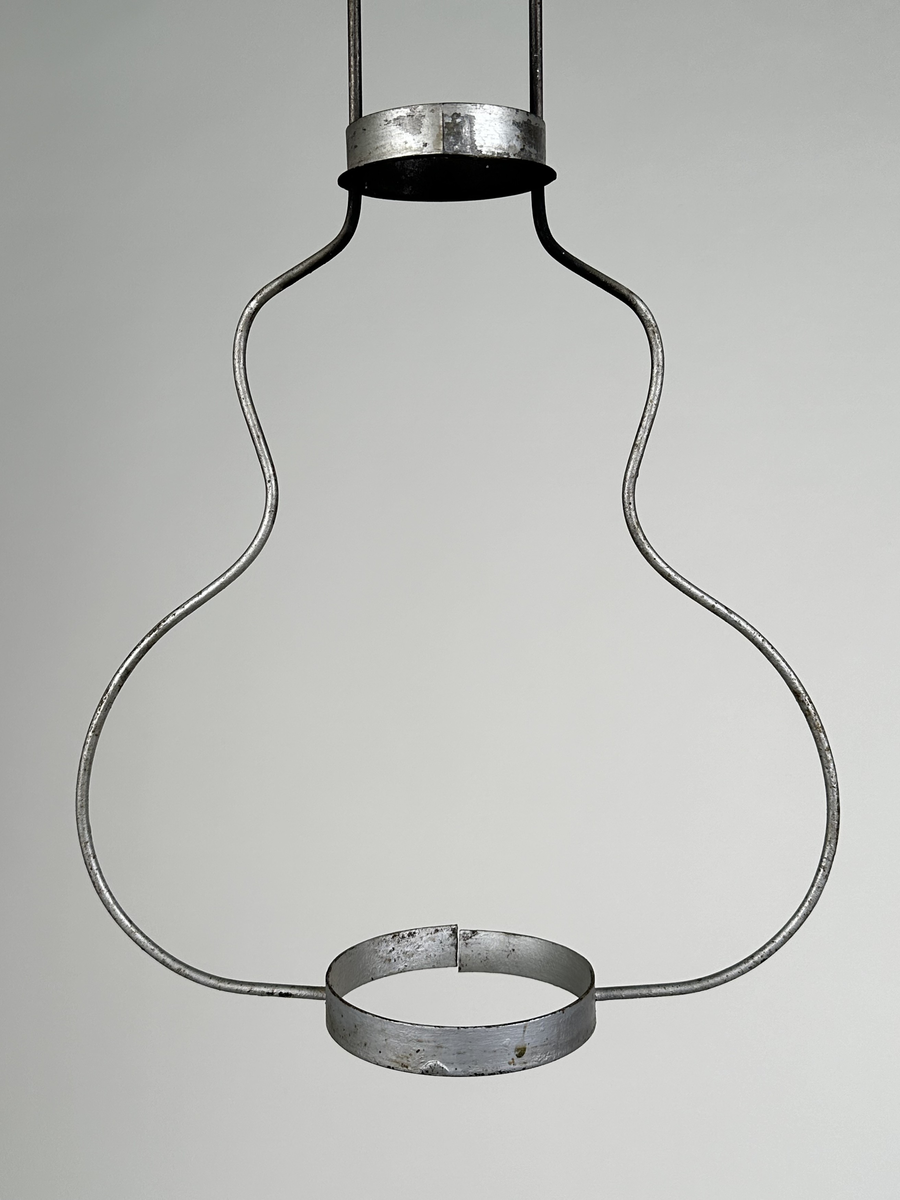 Hängande ställning för fotogenlampa, av två grova metalltrådar som bildar en gitarrform, med ringar av bandplåt nertill samt på halsen för att stödja lampan. Rökhatt med två urtag för metalltrådarna fäst högst upp i ställningen. Silvermålad. Hänger i kätting.