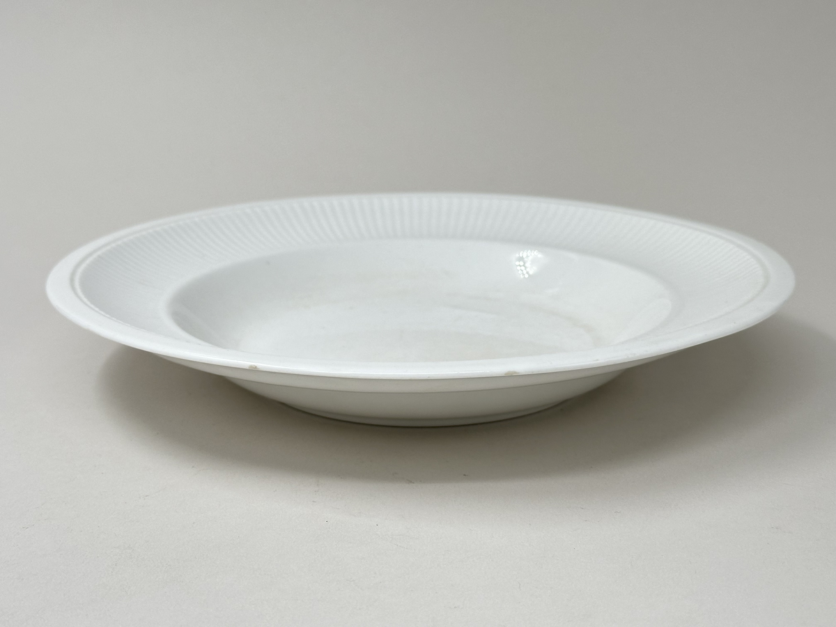 Djup tallrik, av vitglaserad keramik. Räfflat, aningen kupat brätte med slät kant ytterst. Glaserad fotring. Nagg i kanten.
