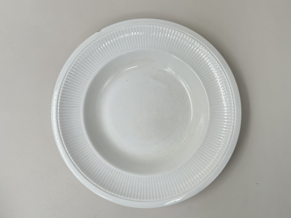 Djup tallrik, av vitglaserad keramik. Räfflat, aningen kupat brätte med slät kant ytterst. Glaserad fotring. Nagg i kanten.