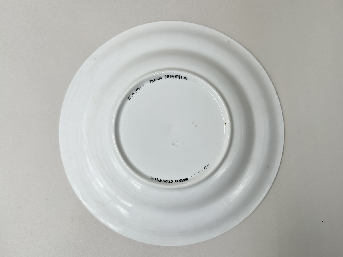 Djup tallrik, av vitglaserad keramik. Räfflat, aningen kupat brätte med slät kant ytterst. Glaserad fotring. Nagg i kanten.