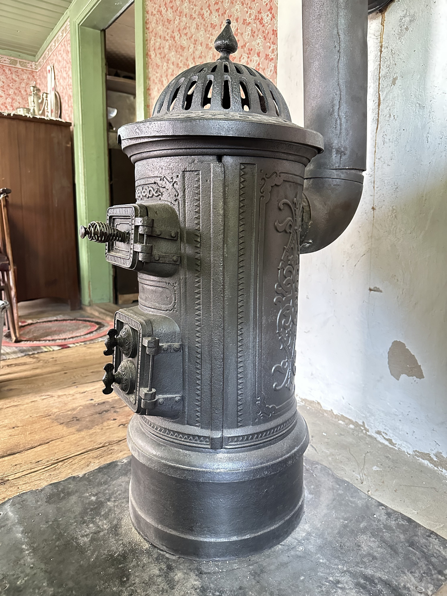 Cylindrisk gjutjärnskamin med spisringar och löstagbar, genombruten krona. Dekor av stiliserade bladrankor i godset. Vedlucka med spiralhandtag, asklucka med två dragventiler reglerade med vingmuttrar. Kaminen kopplad till rund trumma i bakkant. Kronan skadad i kanten. 