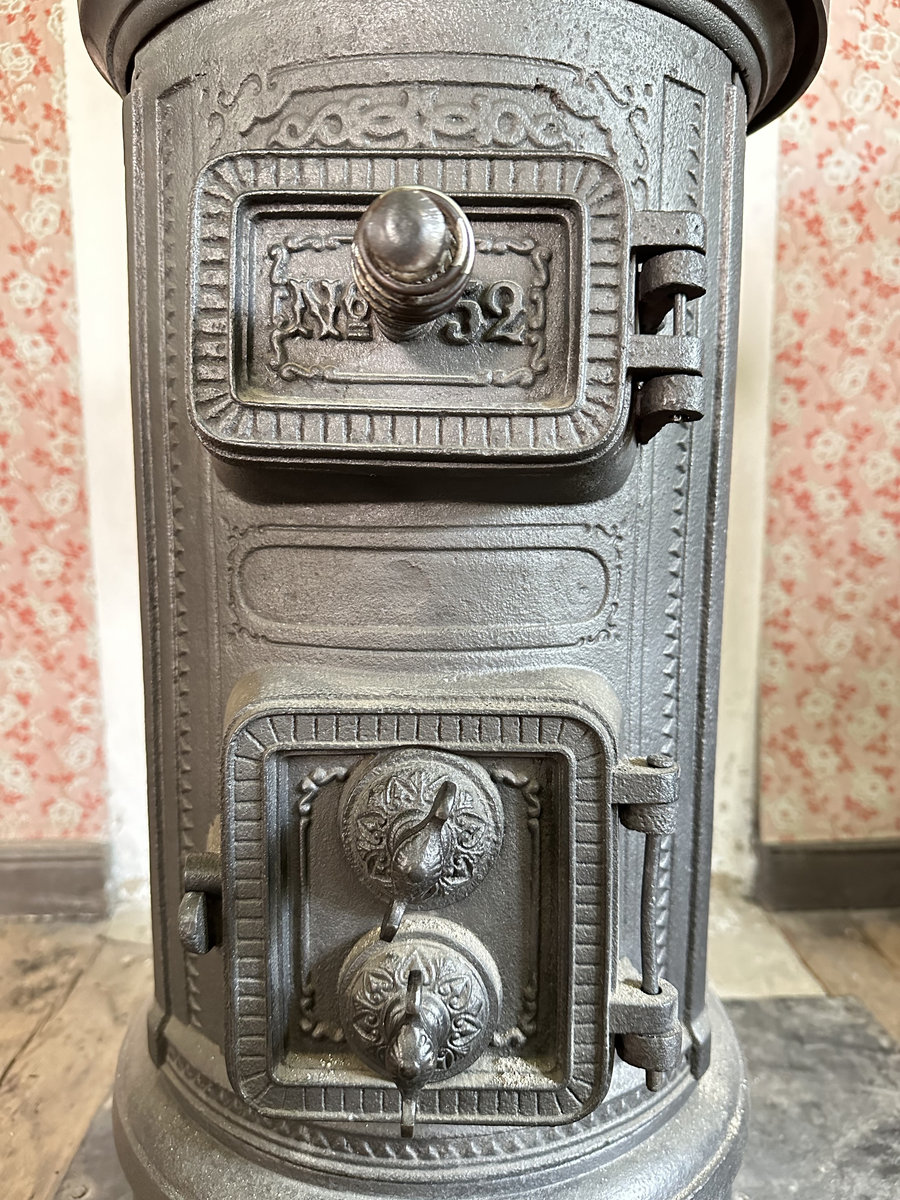 Cylindrisk gjutjärnskamin med spisringar och löstagbar, genombruten krona. Dekor av stiliserade bladrankor i godset. Vedlucka med spiralhandtag, asklucka med två dragventiler reglerade med vingmuttrar. Kaminen kopplad till rund trumma i bakkant. Kronan skadad i kanten. 