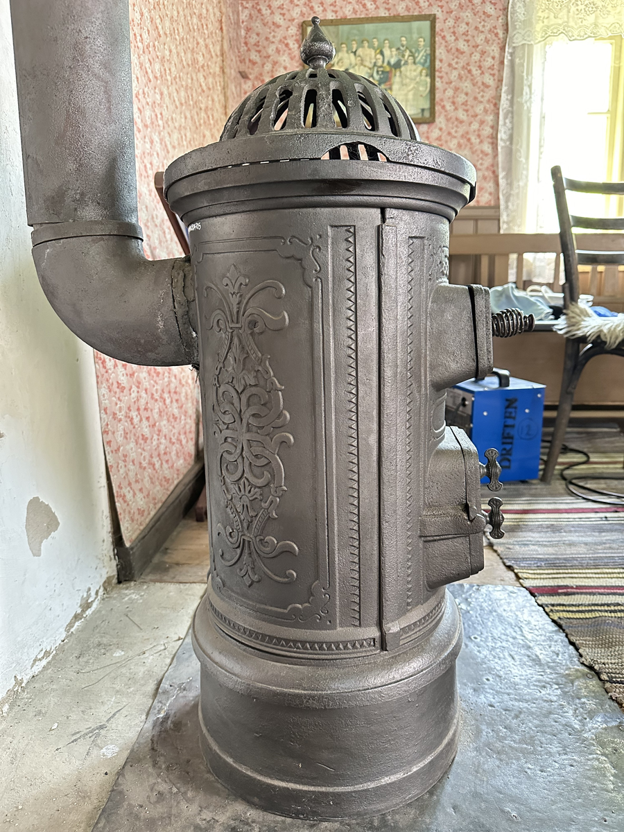 Cylindrisk gjutjärnskamin med spisringar och löstagbar, genombruten krona. Dekor av stiliserade bladrankor i godset. Vedlucka med spiralhandtag, asklucka med två dragventiler reglerade med vingmuttrar. Kaminen kopplad till rund trumma i bakkant. Kronan skadad i kanten. 