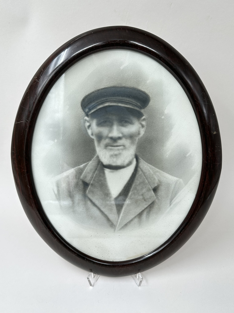 Fotografi i ram. Svart-vit bröstbild av man i skärmmössa och kavaj. Glas och brun träram. Ovalt format. 