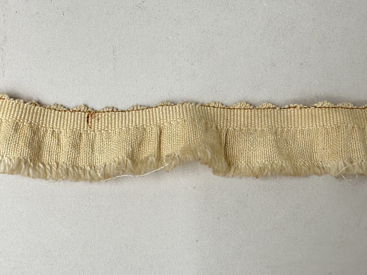 Hyllbård, av ljusgul bomull. Maskinvävt. Uddkant upptill och ett par millimeter frans.