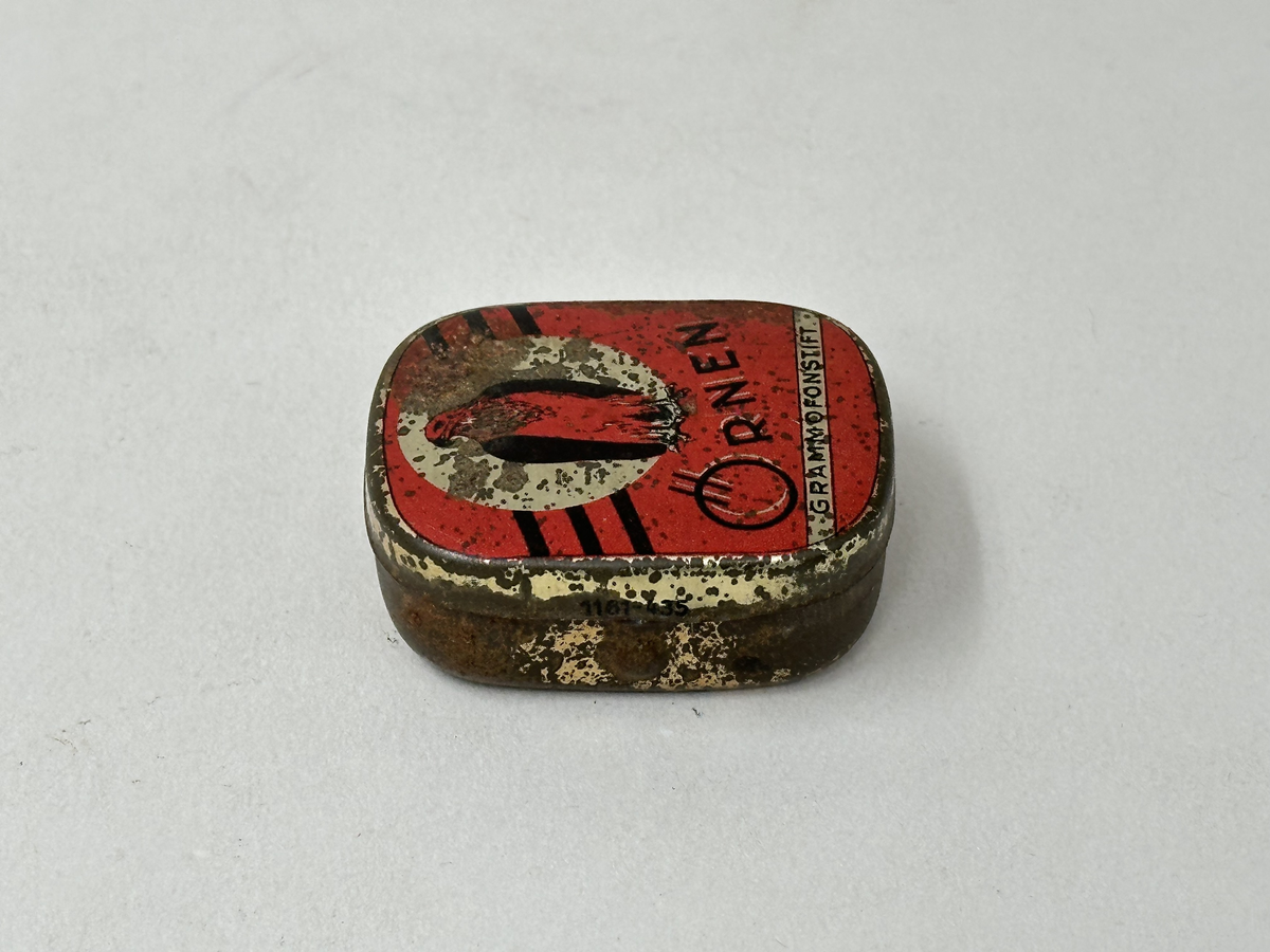 Ask med lock, av plåt. Målad i guld med rött tryck och svart text "ÖRNEN GRAMMOFONSTIFT" på locket. Rostig. Innehållande en liten metallkula och två grammofonstift. 