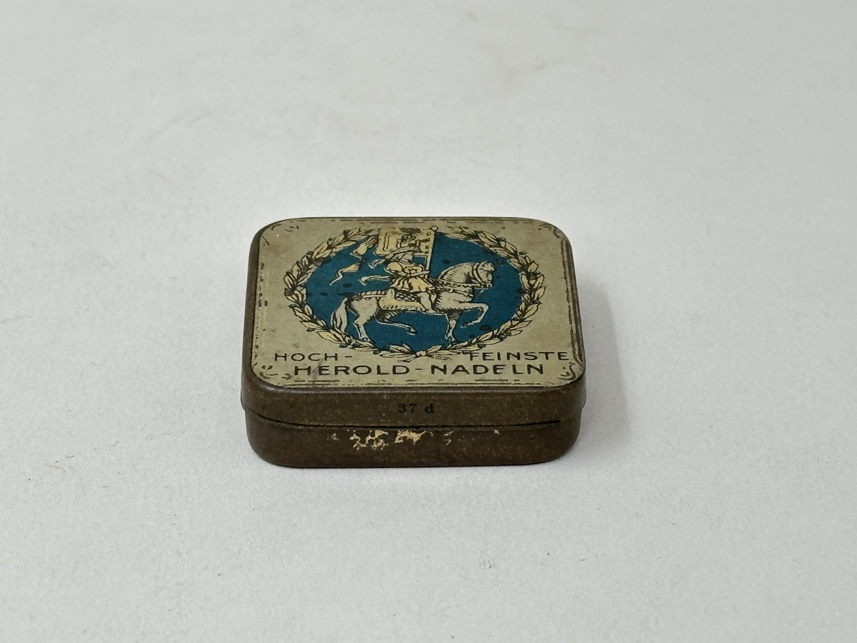 Plåtask med lock. Locket vitt med rund bild: Ryttare med standar mot blå bakgrund, omgiven av bladkrans. Tryckt text: "HOCH-FEINSTE HEROLD-NADELN" "MADE IN GERMANY" "37 D". Innehåller ett skyddspapper och 20 grammofonstift.