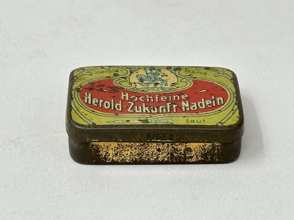 Plåtask med lock, guldmålad. Locket ljusgrönt med tryckt bildrundel: ryttare med standar och text mot röd botten: "Hochfeine Herold-Zukunft-Nadeln". Asken innehåller skyddspapper och grammofonstift.