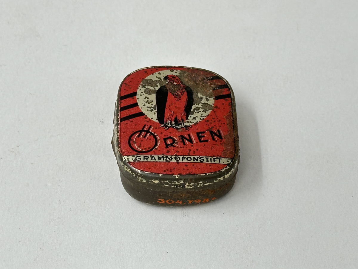 Ask med lock, av plåt. Målad i guld med rött tryck och svart text "ÖRNEN GRAMMOFONSTIFT" på locket. Rostig. Innehållande en liten metallkula och två grammofonstift. 
