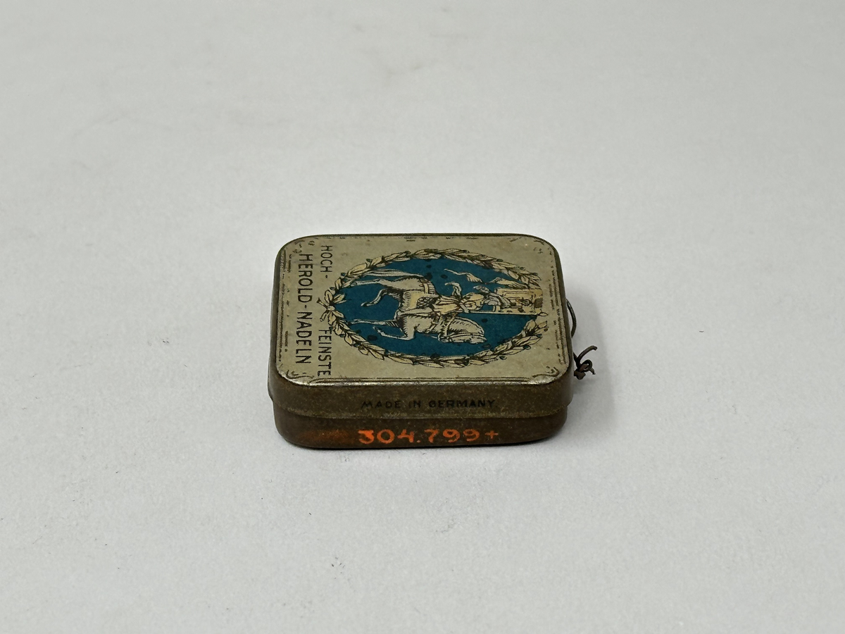 Plåtask med lock. Locket vitt med rund bild: Ryttare med standar mot blå bakgrund, omgiven av bladkrans. Tryckt text: "HOCH-FEINSTE HEROLD-NADELN" "MADE IN GERMANY" "37 D". Innehåller ett skyddspapper och 20 grammofonstift.