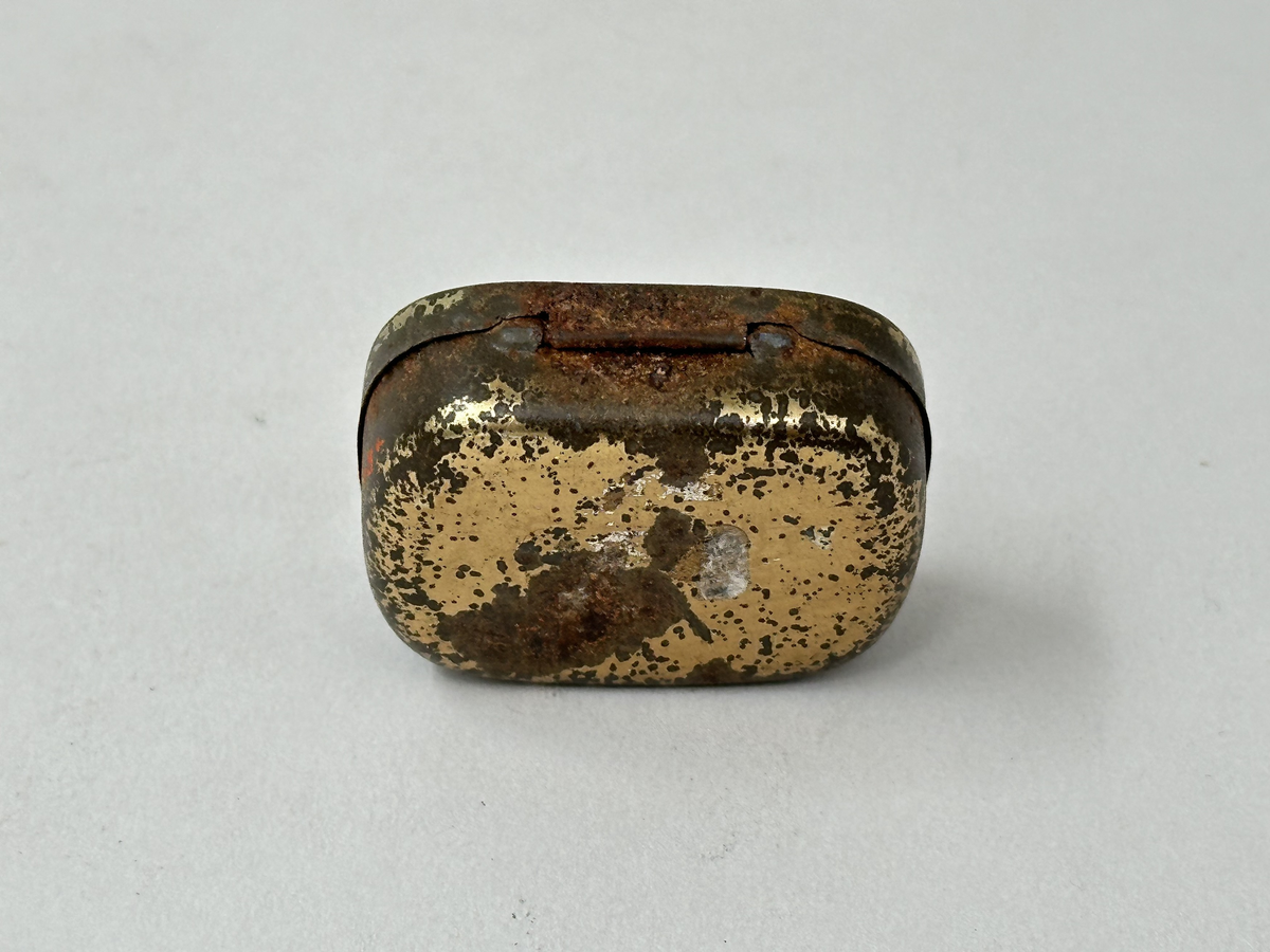 Ask med lock, av plåt. Målad i guld med rött tryck och svart text "ÖRNEN GRAMMOFONSTIFT" på locket. Rostig. Innehållande en liten metallkula och två grammofonstift. 