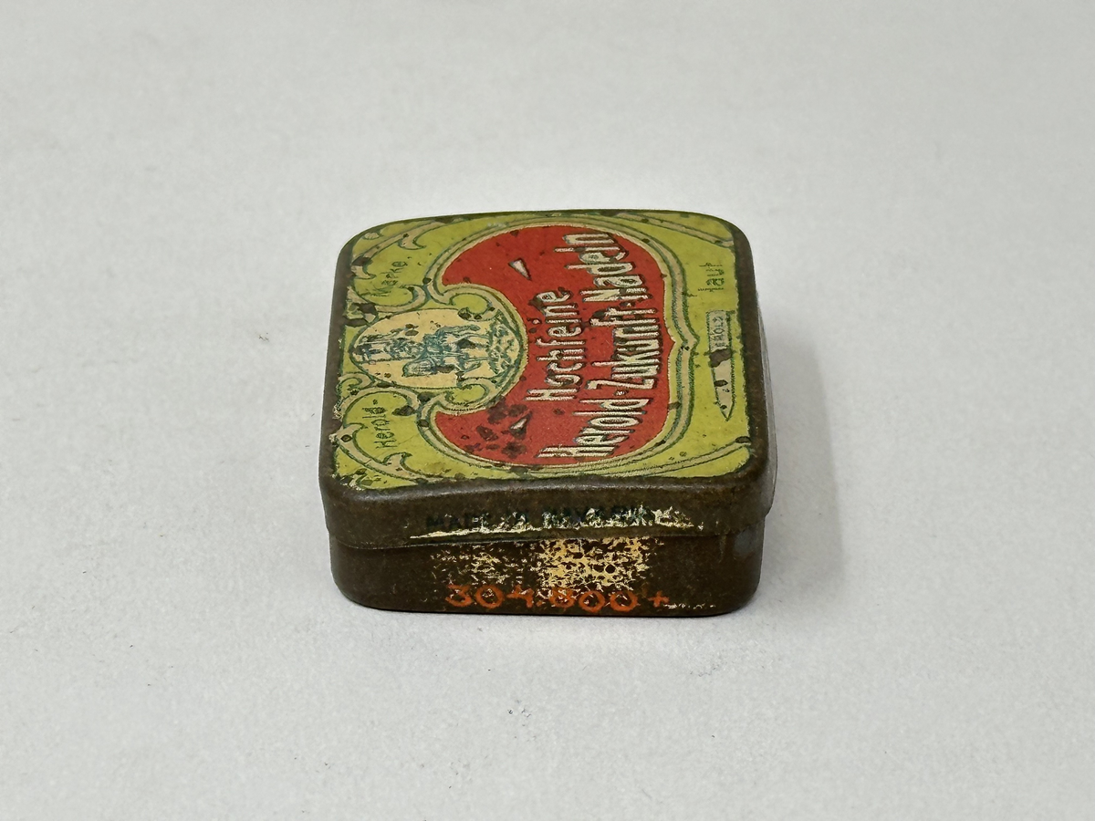 Plåtask med lock, guldmålad. Locket ljusgrönt med tryckt bildrundel: ryttare med standar och text mot röd botten: "Hochfeine Herold-Zukunft-Nadeln". Asken innehåller skyddspapper och grammofonstift.