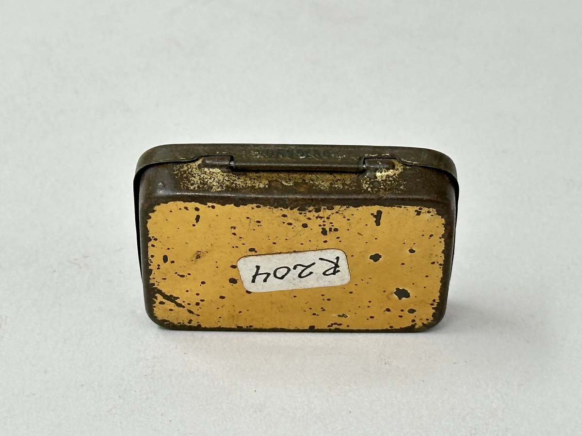 Plåtask med lock, guldmålad. Locket ljusgrönt med tryckt bildrundel: ryttare med standar och text mot röd botten: "Hochfeine Herold-Zukunft-Nadeln". Asken innehåller skyddspapper och grammofonstift.