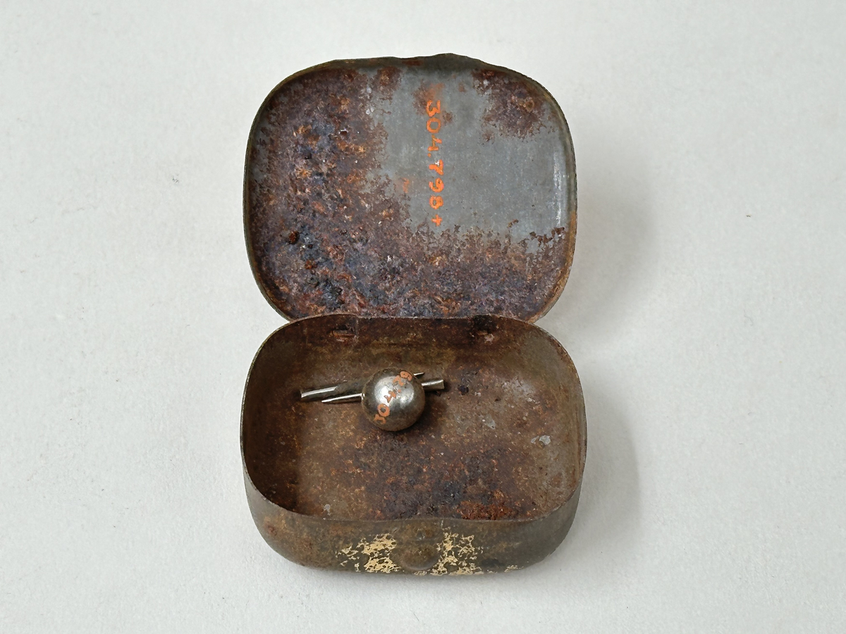 Ask med lock, av plåt. Målad i guld med rött tryck och svart text "ÖRNEN GRAMMOFONSTIFT" på locket. Rostig. Innehållande en liten metallkula och två grammofonstift. 