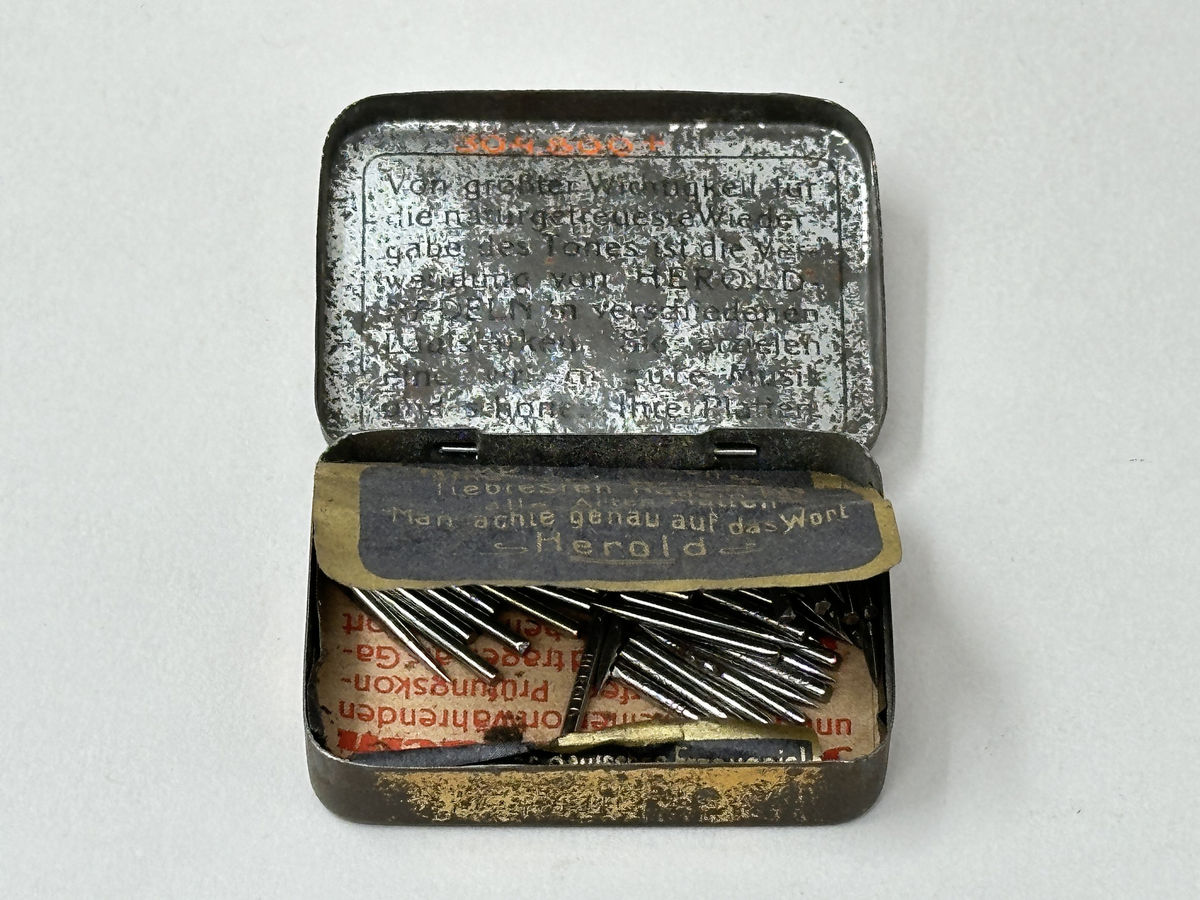 Plåtask med lock, guldmålad. Locket ljusgrönt med tryckt bildrundel: ryttare med standar och text mot röd botten: "Hochfeine Herold-Zukunft-Nadeln". Asken innehåller skyddspapper och grammofonstift.