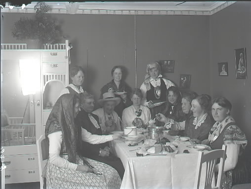 Elva kvinnor sitter vid ett bord varav ett par klädda i hucklen, hatt och/eller folkdräktslik utstyrsel. Man äter soppa, ost och bröd. Bilden är tagen i ett hem, Thorssons hus, med för tiden moderna, ljusa jugendmöbler. Beställare: Fröken Ljungberger.