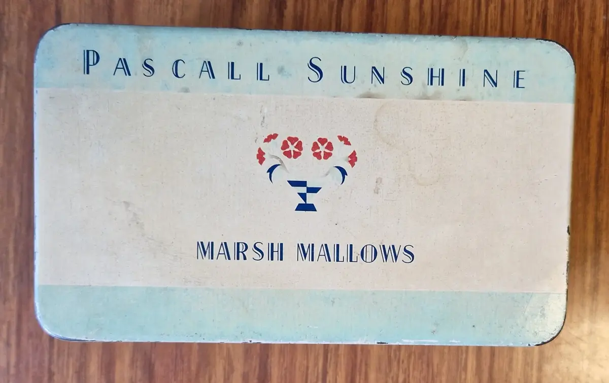 Rektangulär plåtburk i ljusa färger. "Pascall Sunshine. Marsh Mallows" på ovansidan och långsidorna. På kortsidorna "Pascall. Marsh Mallows. Mitcham. Surrey England:".


Anm: Pascalls adress: 100 Blackfriars Road, London, SE1. Också i Mitcham, Surrey.

Den familjeägda firman startades av James Pascall i syfte att tillverka och sälja konfekt.

James Pascall arbetade som agent för Cadbury's innan han startade upp verksamhet med sin bror Alfred i en liten butik på London's Oxford Street 1866.

Till att börja med tillverkade de sötsaker som växtbaserade hostdroppar, godis och hårda franska karameller.

1877 flyttade de till större lokaler i Blackfriars och utvidgade firmans räckvidd.

1897 förstördes deras lokaler i en eldsvåda, startad av en tidigare anställd. Efter eldsvådan flyttade firman till Mitcham, i Surrey.

1922 British Industries Fair Advert for Sweets, Chocolates and Novelties. Also manufacturers of Confectionery and Toys. (Stand No B.8) 
Källa: https://www.gracesguide.co.uk/James_Pascall