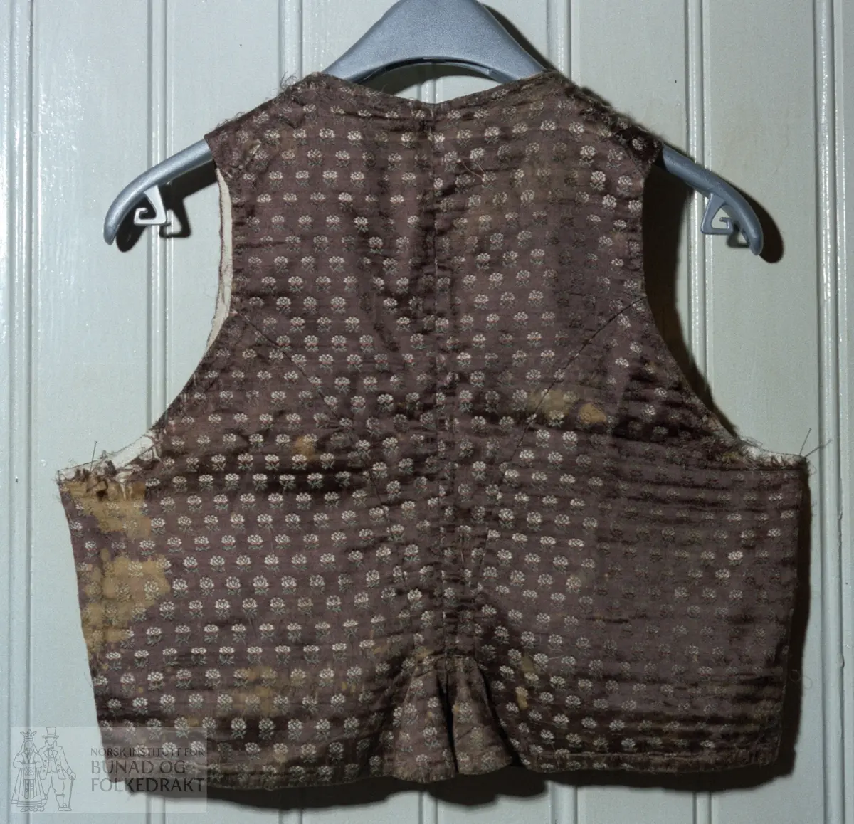 Brun silkedamask med små blomar i kvitt, raudt og grønt.  Fôr: linlerret.  Spilesaum.  2 rader med prikkesting.  Hel lengde: 40 cm.