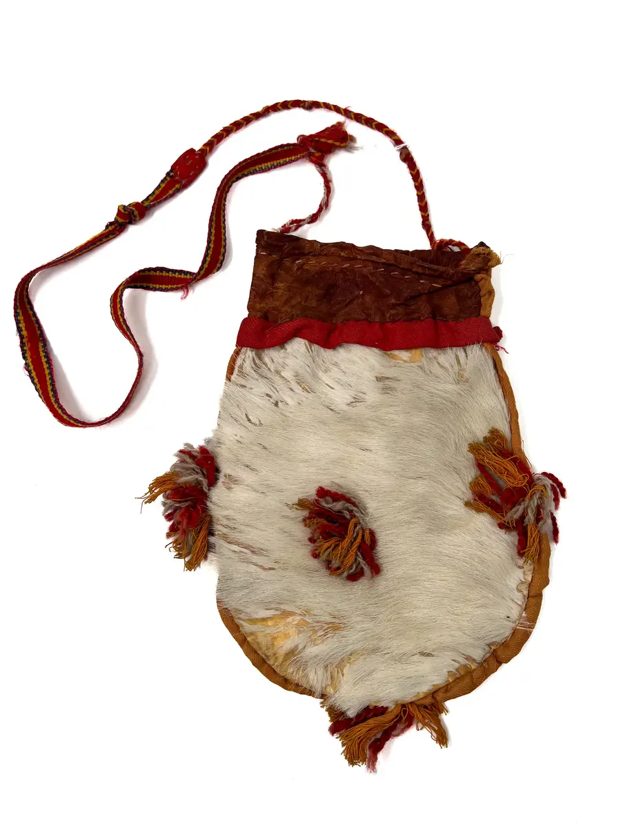 Beskrivning: Väska. Handsydd. Överdelen av garvat skinn. Nederdelen av två sidostycken. Sidostyckena av rundskurna stycken av getpälsskinn. L. 21,5 x B. 18 cm. Mellanlägg av dubbel, ljus, orangefärgad bomullskypert i sömmen mellan de två sidostyckena. Tofsar av grått och rostfärgat, tvåtrådigt ullgarn och orangefärgat bomullsgarn fästade i mitten på sidostyckena, mitt i var sidsöm och mitt i bottensömmen. Överdelen skuren i ett stycke med mellanlägg av ljus, orangefärgad bomullskypert i sidsömmen. I sömmen mellan överdelen och nederdelen mellanlägg av dubbelvikt, 1,5 cm bred, rostfärgad bomullkypert. Kanten på överdelen nervikt mot avigsidan och sydd till en dragsko med öppning åt endast ett håll. Ett snöre i dragskon, vars ändar antagligen är uppätna av djur och därför ej går att beskriva. Upphängningsband vävt av rött, gult och klarblått ullgarn i varpen och lila ullgarn som inslagstråd. En snodd fläta av rött och gult ullgarn i fyrflätning som bildar ett randigt mönster fastsydd i ena änden på bandet. En snodd flätat av gultrött och lila ullgarn, flätat i fiskbenmönster, fästat i bandets andra ände och fastsydd i överdelens sidosöm. Fästena mellan bandet och snoddarna dolda av rött kläde. Den fyrflätade snodden är fastknuten i en snodd av tvinnat, grått och rostfärgat ullgarn, som i sin tur är fastsydd i överdelens andra sida. Väskan är helt och hållet sydd med bomullstråd. 
