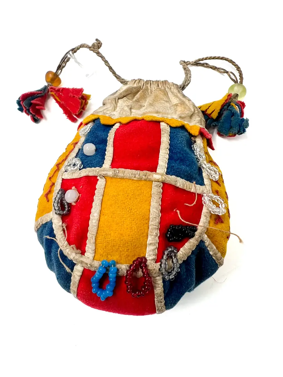  Pung, av kläde och vitt skinn. Handsydd. Överdelen av vitt skinn. Sidorna rundskurna, sydda av bitar av gulrött och klarblått kläde. Bottenstycke av ytterst gult, därefter klarblått och i mitten rött kläde med mellanlägg av dubbelvikt, vitt skinn i sömmarna. Samma typ av mellanlägg i sömmarna mellan sidostyckena och bottenremsan. 

Det ena sidostycket med vita glaspärlor i mitten på varje klädesbit och 15 st öglor gjorda av små glaspärlor, fästade längs kanten på sidostycket. På det andra sidostycket glaspärlor på endast två klädesbitar och 10 st öglor av pärltråd längs kanten. 

Överdel av vitt skinn, sydd av dubbelt skinn med dragsko upptill, dragskon öppen åt båda sidor, genom dragskon två snören, tvinnade av smala skinnremmar med tofsar av klarblått, rött och gult kläde. 

Dragsnörena hålls ihop parvis av en glaspärla, varigenom de är trädda. Sömmarna sydda med bomullstråd och något slags vaxad tråd.