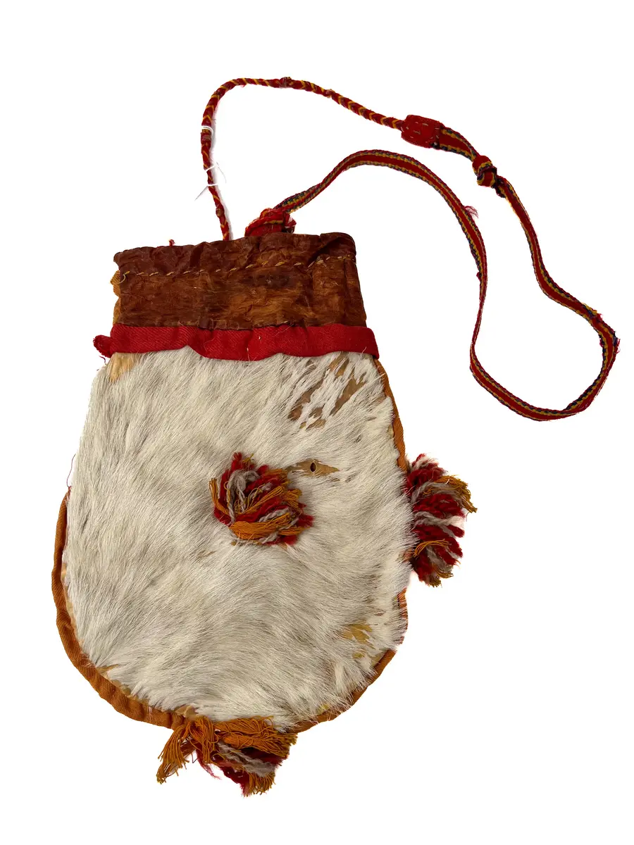 Beskrivning: Väska. Handsydd. Överdelen av garvat skinn. Nederdelen av två sidostycken. Sidostyckena av rundskurna stycken av getpälsskinn. L. 21,5 x B. 18 cm. Mellanlägg av dubbel, ljus, orangefärgad bomullskypert i sömmen mellan de två sidostyckena. Tofsar av grått och rostfärgat, tvåtrådigt ullgarn och orangefärgat bomullsgarn fästade i mitten på sidostyckena, mitt i var sidsöm och mitt i bottensömmen. Överdelen skuren i ett stycke med mellanlägg av ljus, orangefärgad bomullskypert i sidsömmen. I sömmen mellan överdelen och nederdelen mellanlägg av dubbelvikt, 1,5 cm bred, rostfärgad bomullkypert. Kanten på överdelen nervikt mot avigsidan och sydd till en dragsko med öppning åt endast ett håll. Ett snöre i dragskon, vars ändar antagligen är uppätna av djur och därför ej går att beskriva. Upphängningsband vävt av rött, gult och klarblått ullgarn i varpen och lila ullgarn som inslagstråd. En snodd fläta av rött och gult ullgarn i fyrflätning som bildar ett randigt mönster fastsydd i ena änden på bandet. En snodd flätat av gultrött och lila ullgarn, flätat i fiskbenmönster, fästat i bandets andra ände och fastsydd i överdelens sidosöm. Fästena mellan bandet och snoddarna dolda av rött kläde. Den fyrflätade snodden är fastknuten i en snodd av tvinnat, grått och rostfärgat ullgarn, som i sin tur är fastsydd i överdelens andra sida. Väskan är helt och hållet sydd med bomullstråd. 