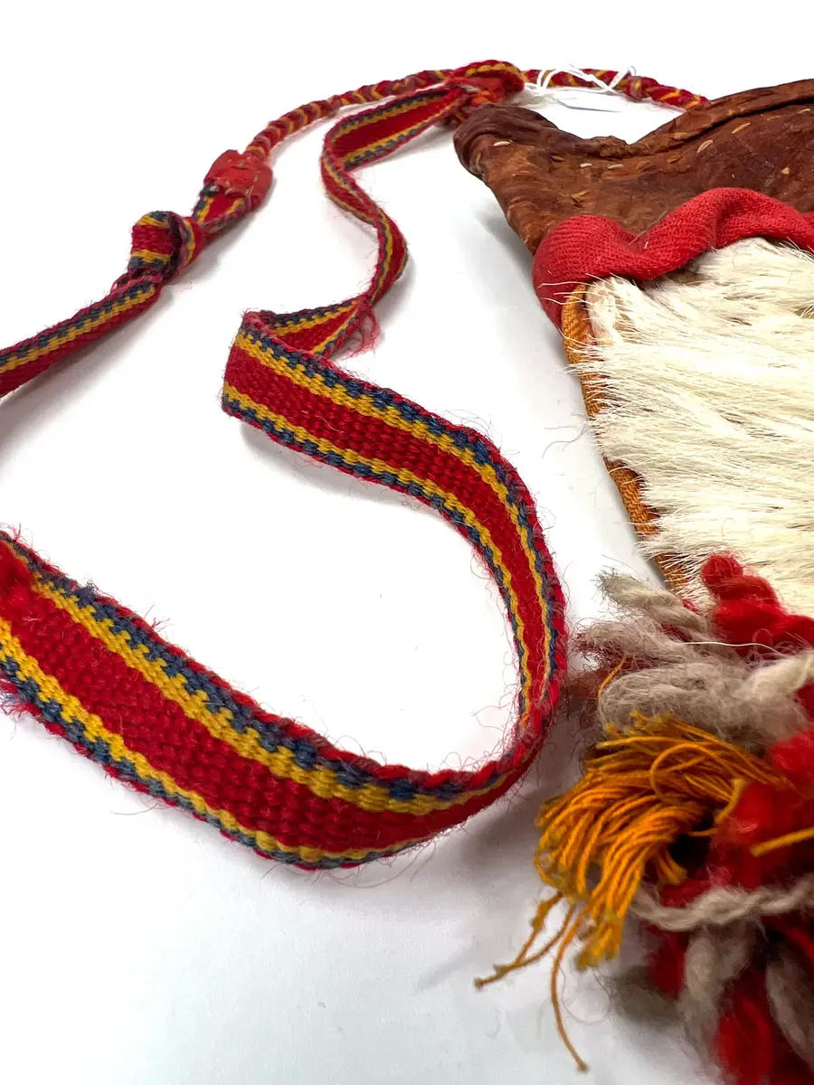 Beskrivning: Väska. Handsydd. Överdelen av garvat skinn. Nederdelen av två sidostycken. Sidostyckena av rundskurna stycken av getpälsskinn. L. 21,5 x B. 18 cm. Mellanlägg av dubbel, ljus, orangefärgad bomullskypert i sömmen mellan de två sidostyckena. Tofsar av grått och rostfärgat, tvåtrådigt ullgarn och orangefärgat bomullsgarn fästade i mitten på sidostyckena, mitt i var sidsöm och mitt i bottensömmen. Överdelen skuren i ett stycke med mellanlägg av ljus, orangefärgad bomullskypert i sidsömmen. I sömmen mellan överdelen och nederdelen mellanlägg av dubbelvikt, 1,5 cm bred, rostfärgad bomullkypert. Kanten på överdelen nervikt mot avigsidan och sydd till en dragsko med öppning åt endast ett håll. Ett snöre i dragskon, vars ändar antagligen är uppätna av djur och därför ej går att beskriva. Upphängningsband vävt av rött, gult och klarblått ullgarn i varpen och lila ullgarn som inslagstråd. En snodd fläta av rött och gult ullgarn i fyrflätning som bildar ett randigt mönster fastsydd i ena änden på bandet. En snodd flätat av gultrött och lila ullgarn, flätat i fiskbenmönster, fästat i bandets andra ände och fastsydd i överdelens sidosöm. Fästena mellan bandet och snoddarna dolda av rött kläde. Den fyrflätade snodden är fastknuten i en snodd av tvinnat, grått och rostfärgat ullgarn, som i sin tur är fastsydd i överdelens andra sida. Väskan är helt och hållet sydd med bomullstråd. 