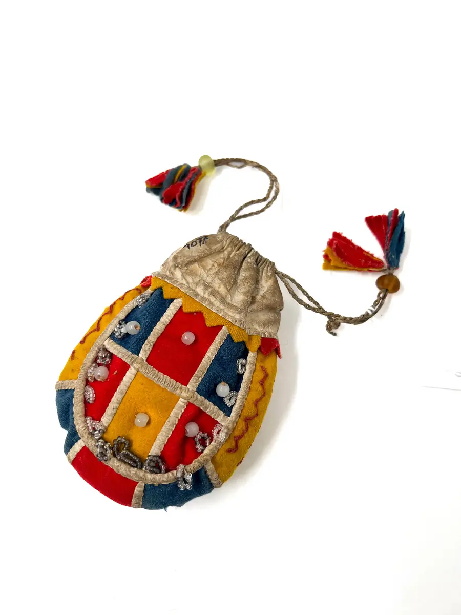  Pung, av kläde och vitt skinn. Handsydd. Överdelen av vitt skinn. Sidorna rundskurna, sydda av bitar av gulrött och klarblått kläde. Bottenstycke av ytterst gult, därefter klarblått och i mitten rött kläde med mellanlägg av dubbelvikt, vitt skinn i sömmarna. Samma typ av mellanlägg i sömmarna mellan sidostyckena och bottenremsan. 

Det ena sidostycket med vita glaspärlor i mitten på varje klädesbit och 15 st öglor gjorda av små glaspärlor, fästade längs kanten på sidostycket. På det andra sidostycket glaspärlor på endast två klädesbitar och 10 st öglor av pärltråd längs kanten. 

Överdel av vitt skinn, sydd av dubbelt skinn med dragsko upptill, dragskon öppen åt båda sidor, genom dragskon två snören, tvinnade av smala skinnremmar med tofsar av klarblått, rött och gult kläde. 

Dragsnörena hålls ihop parvis av en glaspärla, varigenom de är trädda. Sömmarna sydda med bomullstråd och något slags vaxad tråd.