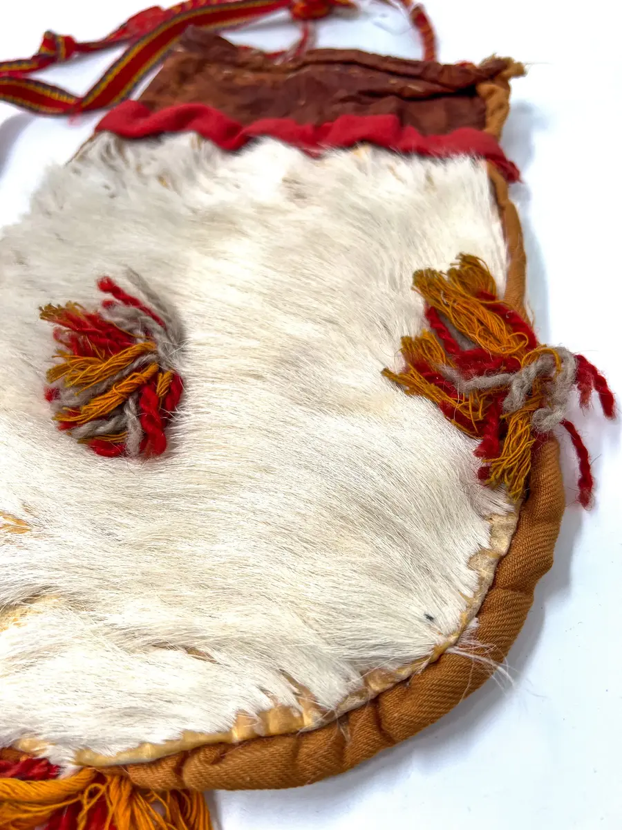 Beskrivning: Väska. Handsydd. Överdelen av garvat skinn. Nederdelen av två sidostycken. Sidostyckena av rundskurna stycken av getpälsskinn. L. 21,5 x B. 18 cm. Mellanlägg av dubbel, ljus, orangefärgad bomullskypert i sömmen mellan de två sidostyckena. Tofsar av grått och rostfärgat, tvåtrådigt ullgarn och orangefärgat bomullsgarn fästade i mitten på sidostyckena, mitt i var sidsöm och mitt i bottensömmen. Överdelen skuren i ett stycke med mellanlägg av ljus, orangefärgad bomullskypert i sidsömmen. I sömmen mellan överdelen och nederdelen mellanlägg av dubbelvikt, 1,5 cm bred, rostfärgad bomullkypert. Kanten på överdelen nervikt mot avigsidan och sydd till en dragsko med öppning åt endast ett håll. Ett snöre i dragskon, vars ändar antagligen är uppätna av djur och därför ej går att beskriva. Upphängningsband vävt av rött, gult och klarblått ullgarn i varpen och lila ullgarn som inslagstråd. En snodd fläta av rött och gult ullgarn i fyrflätning som bildar ett randigt mönster fastsydd i ena änden på bandet. En snodd flätat av gultrött och lila ullgarn, flätat i fiskbenmönster, fästat i bandets andra ände och fastsydd i överdelens sidosöm. Fästena mellan bandet och snoddarna dolda av rött kläde. Den fyrflätade snodden är fastknuten i en snodd av tvinnat, grått och rostfärgat ullgarn, som i sin tur är fastsydd i överdelens andra sida. Väskan är helt och hållet sydd med bomullstråd. 