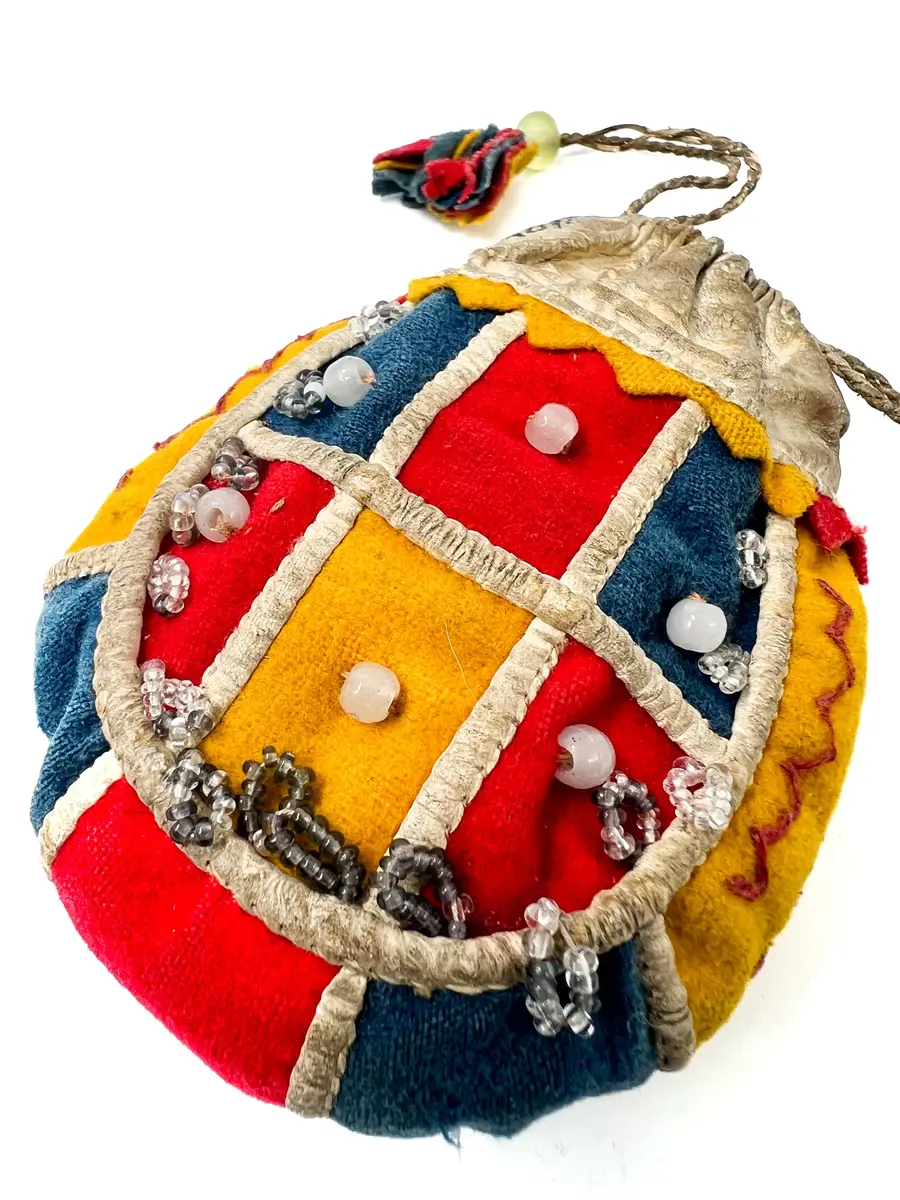  Pung, av kläde och vitt skinn. Handsydd. Överdelen av vitt skinn. Sidorna rundskurna, sydda av bitar av gulrött och klarblått kläde. Bottenstycke av ytterst gult, därefter klarblått och i mitten rött kläde med mellanlägg av dubbelvikt, vitt skinn i sömmarna. Samma typ av mellanlägg i sömmarna mellan sidostyckena och bottenremsan. 

Det ena sidostycket med vita glaspärlor i mitten på varje klädesbit och 15 st öglor gjorda av små glaspärlor, fästade längs kanten på sidostycket. På det andra sidostycket glaspärlor på endast två klädesbitar och 10 st öglor av pärltråd längs kanten. 

Överdel av vitt skinn, sydd av dubbelt skinn med dragsko upptill, dragskon öppen åt båda sidor, genom dragskon två snören, tvinnade av smala skinnremmar med tofsar av klarblått, rött och gult kläde. 

Dragsnörena hålls ihop parvis av en glaspärla, varigenom de är trädda. Sömmarna sydda med bomullstråd och något slags vaxad tråd.