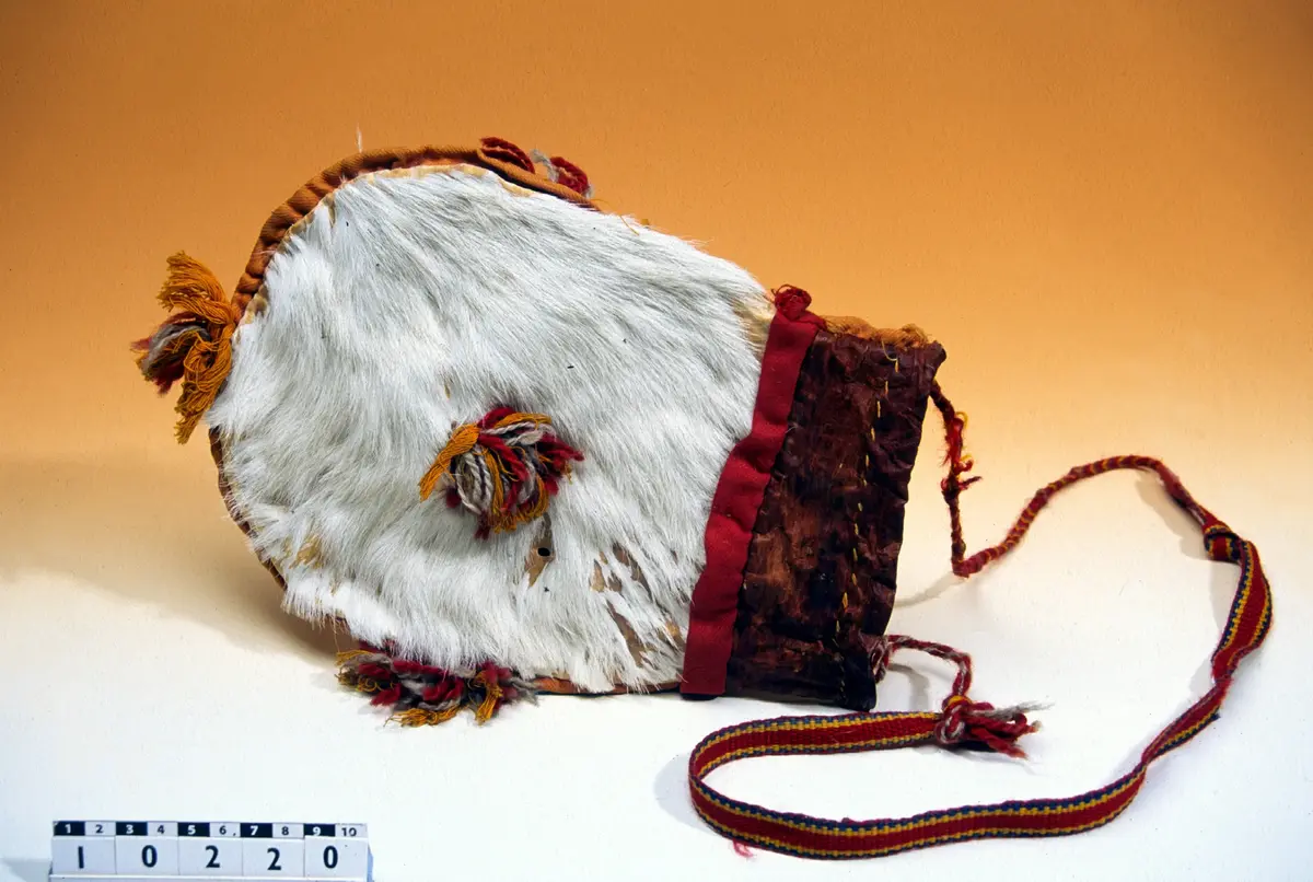 Beskrivning: Väska. Handsydd. Överdelen av garvat skinn. Nederdelen av två sidostycken. Sidostyckena av rundskurna stycken av getpälsskinn. L. 21,5 x B. 18 cm. Mellanlägg av dubbel, ljus, orangefärgad bomullskypert i sömmen mellan de två sidostyckena. Tofsar av grått och rostfärgat, tvåtrådigt ullgarn och orangefärgat bomullsgarn fästade i mitten på sidostyckena, mitt i var sidsöm och mitt i bottensömmen. Överdelen skuren i ett stycke med mellanlägg av ljus, orangefärgad bomullskypert i sidsömmen. I sömmen mellan överdelen och nederdelen mellanlägg av dubbelvikt, 1,5 cm bred, rostfärgad bomullkypert. Kanten på överdelen nervikt mot avigsidan och sydd till en dragsko med öppning åt endast ett håll. Ett snöre i dragskon, vars ändar antagligen är uppätna av djur och därför ej går att beskriva. Upphängningsband vävt av rött, gult och klarblått ullgarn i varpen och lila ullgarn som inslagstråd. En snodd fläta av rött och gult ullgarn i fyrflätning som bildar ett randigt mönster fastsydd i ena änden på bandet. En snodd flätat av gultrött och lila ullgarn, flätat i fiskbenmönster, fästat i bandets andra ände och fastsydd i överdelens sidosöm. Fästena mellan bandet och snoddarna dolda av rött kläde. Den fyrflätade snodden är fastknuten i en snodd av tvinnat, grått och rostfärgat ullgarn, som i sin tur är fastsydd i överdelens andra sida. Väskan är helt och hållet sydd med bomullstråd. 