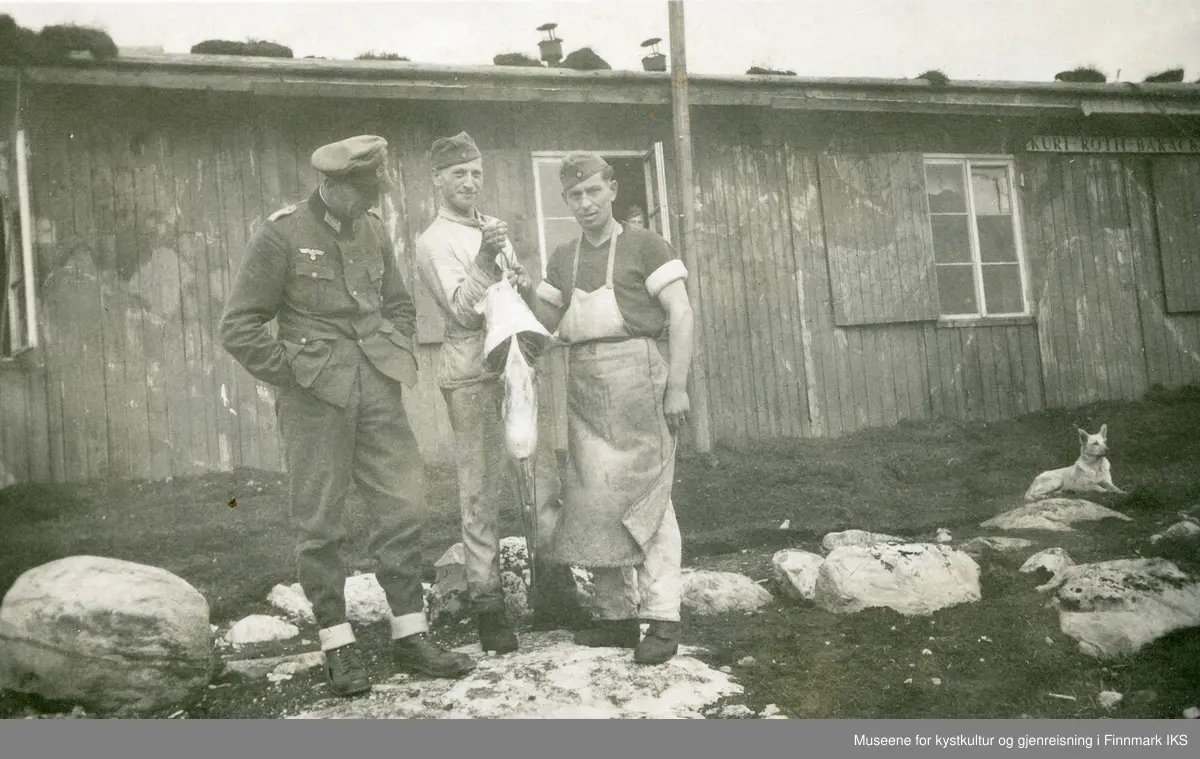 Tre tyske soldater foran en brakke. To av dem holder opp en stor fisk ...