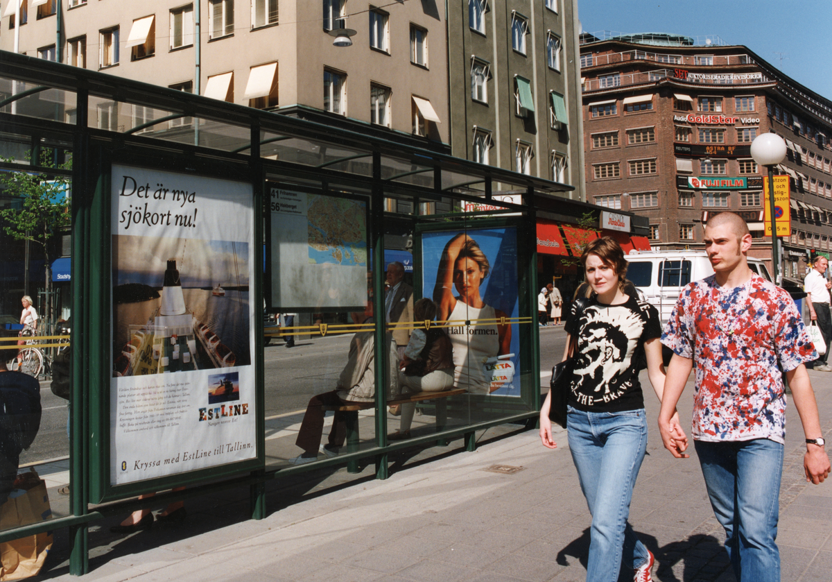 Reklamskylt för Estline i en busskur/busshållplats på Kungsgatan. Människor flanerar och bilar trafikerar gatan.