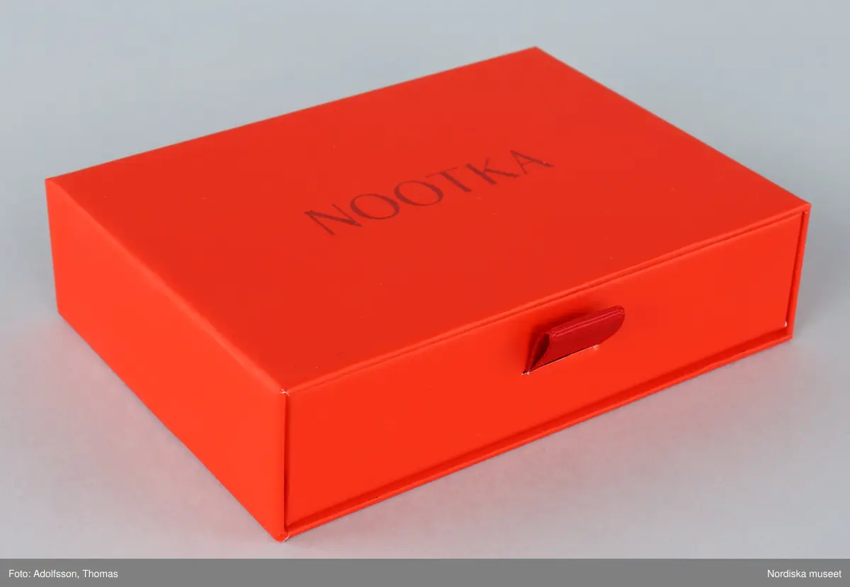 Förpackning för örhängen.
a-b) Ask av orange papp, rektangulär, med låda inskjuten från ena långsidan. Asken med handtag av tygband. Text på asken "NOOTKA".
c-d) Två påsar av orange filtat syntetmaterial. Rektangulära med flik vikt över öppningen. Text på framsidan "NOOTKA".