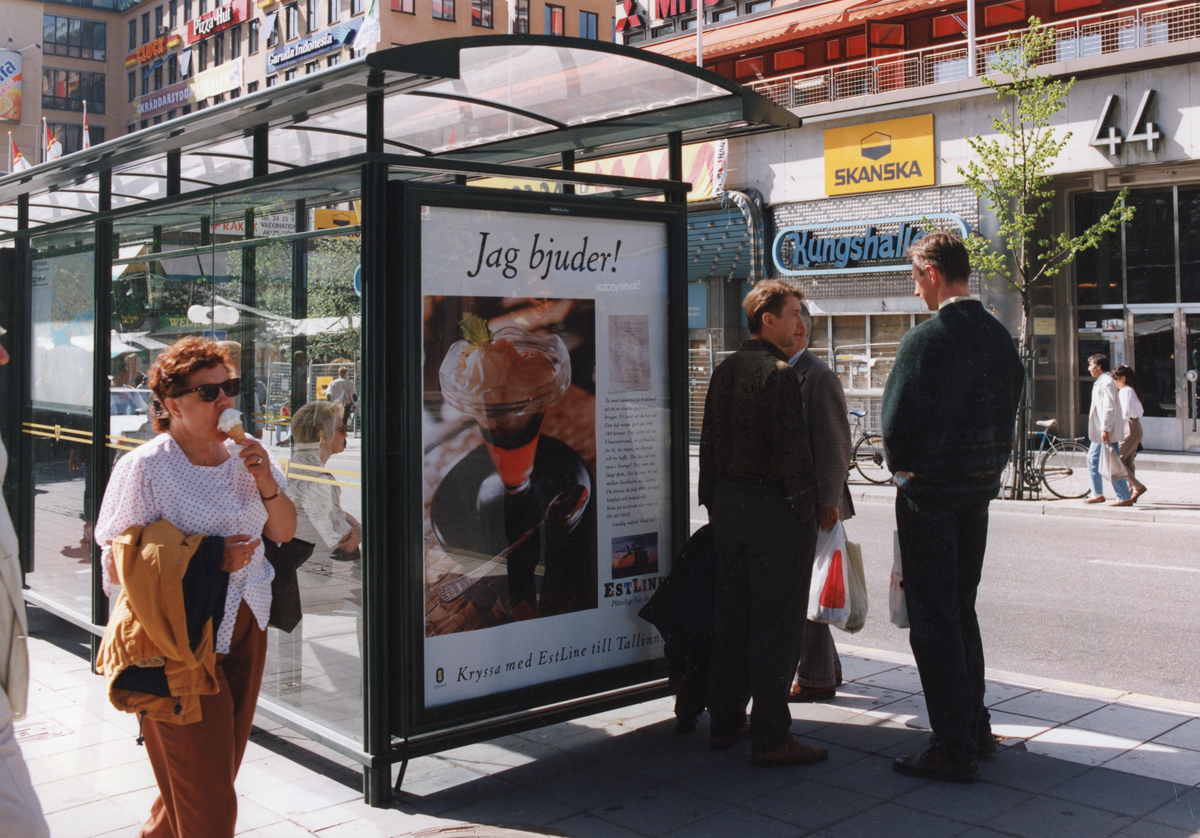 Reklamaffisch för Estline på busskur/busshållplats på Kungsgatan. Människor väntar på bussen och en kvinna äter glass.