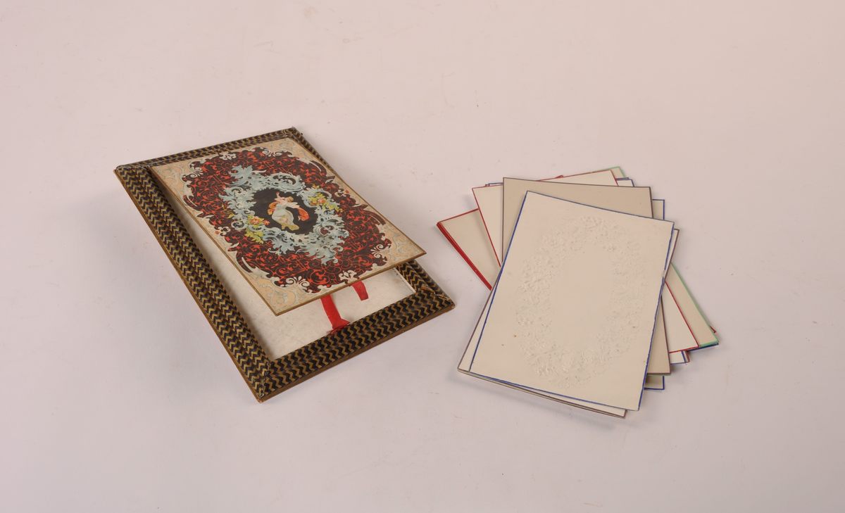 Minnebok i presset papp. Hengslet lokk med ornamentikk i hvitt, blått, rødt og grønt, som omslutter et sort felt med en flyvende engel. Løse ubeskrevne ark innvendig, med farget kant. Topparket er preget.