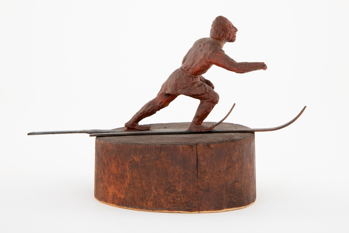 Modell i rødbrun voks av skiløper, på ski av kobber. Figuren står på en rund treblokk, malt eller beiset rødbrunt. Modell til NK 3103 (i sølv).