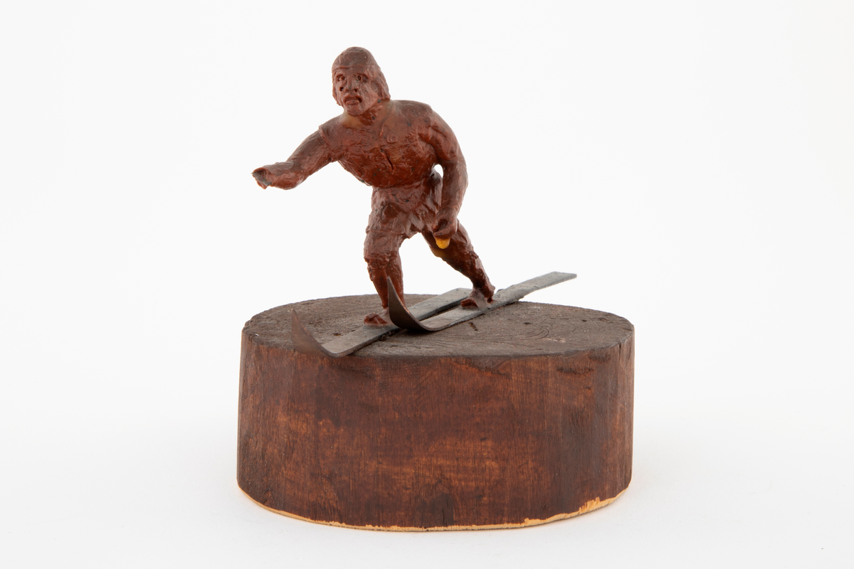 Modell i rødbrun voks av skiløper, på ski av kobber. Figuren står på en rund treblokk, malt eller beiset rødbrunt. Modell til NK 3103 (i sølv).