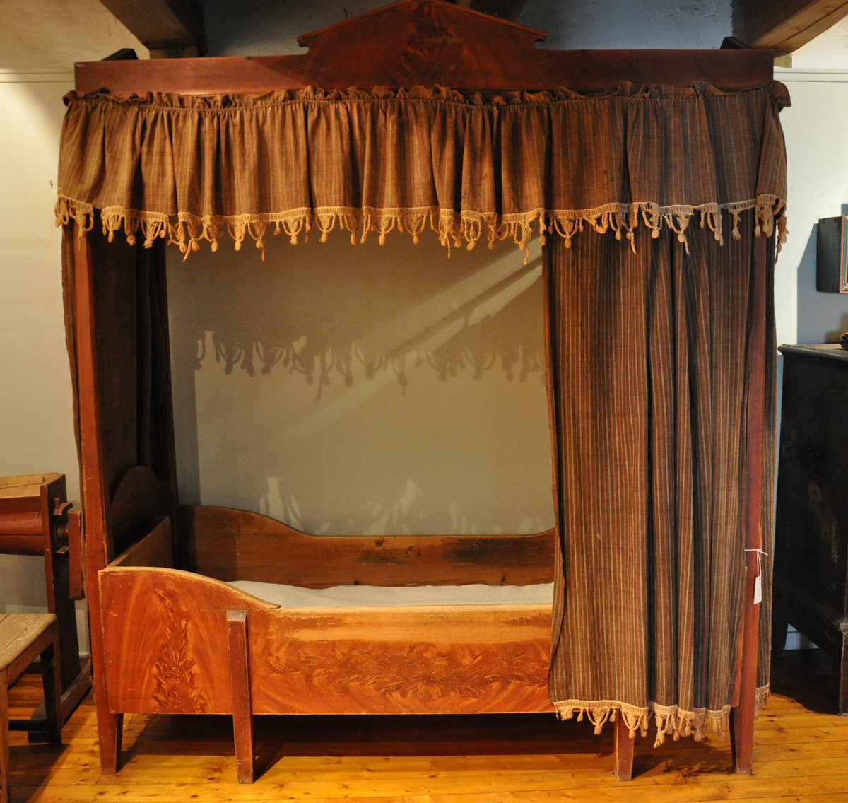 Rødmalt himmelseng med forheng og kappe i tekstil med røde, hvite og lyseblå striper på brun bakgrunn, med påsydd dekorativ hvit kant med possementer. Toppstykket og sidestykke ådret som mahogni.