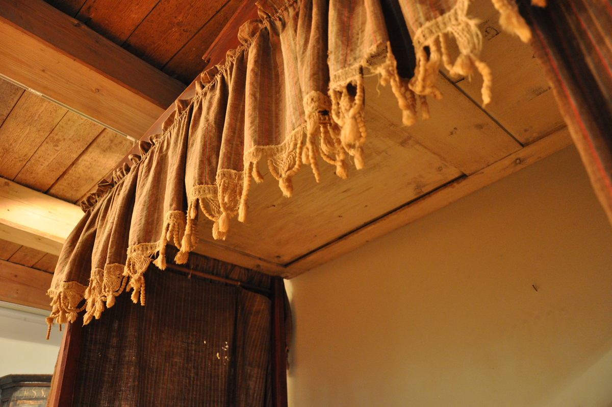 Rødmalt himmelseng med forheng og kappe i tekstil med røde, hvite og lyseblå striper på brun bakgrunn, med påsydd dekorativ hvit kant med possementer. Toppstykket og sidestykke ådret som mahogni.