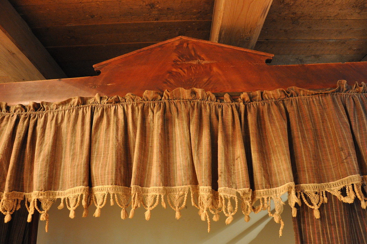 Rødmalt himmelseng med forheng og kappe i tekstil med røde, hvite og lyseblå striper på brun bakgrunn, med påsydd dekorativ hvit kant med possementer. Toppstykket og sidestykke ådret som mahogni.