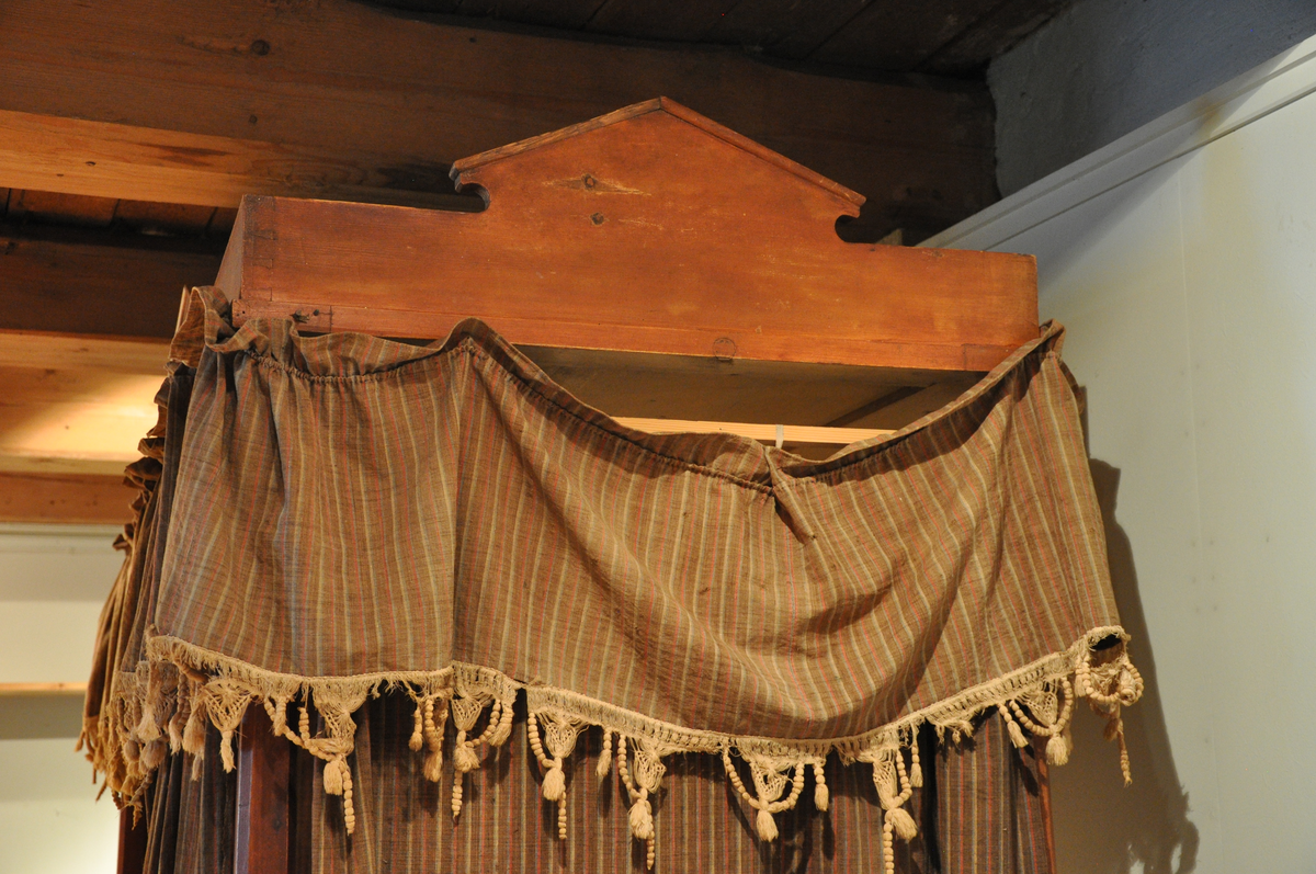 Rødmalt himmelseng med forheng og kappe i tekstil med røde, hvite og lyseblå striper på brun bakgrunn, med påsydd dekorativ hvit kant med possementer. Toppstykket og sidestykke ådret som mahogni.