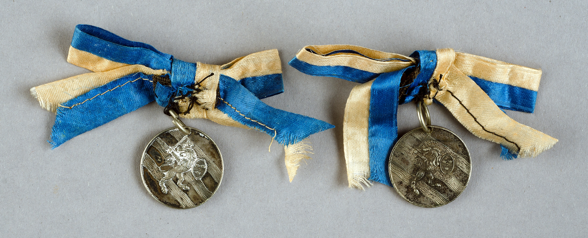Två "medaljonger", av vitmetall, runda med Göteborgs Nations vapen på ena sidan och text "GÖTEBORGS NATION 27/3 1882" på andra sidan, med blågul sidenrosett. 



