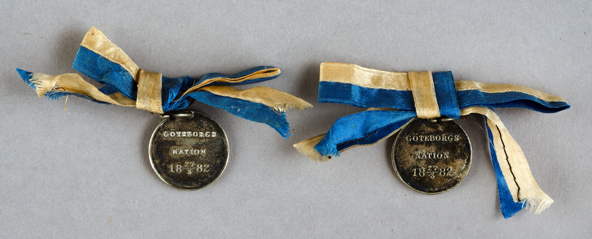 Två "medaljonger", av vitmetall, runda med Göteborgs Nations vapen på ena sidan och text "GÖTEBORGS NATION 27/3 1882" på andra sidan, med blågul sidenrosett. 



