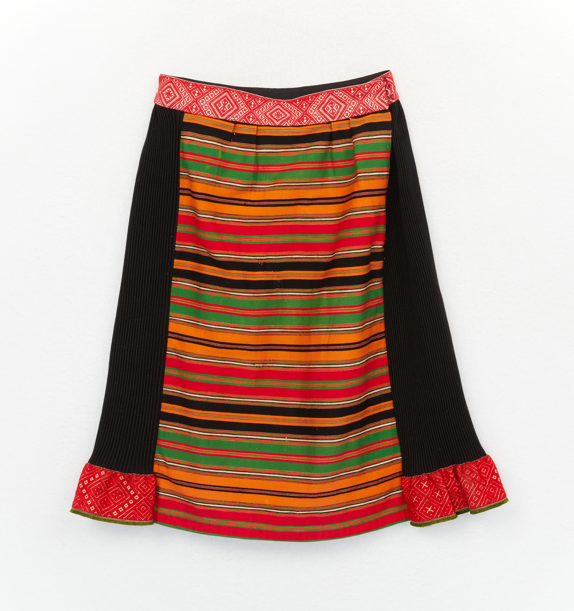 Kjol till Boda sockendräkt för kvinna. Rynkkjol av svart halvylletyg. Bredda av rött, grönt, svart och vitt tvärrandigt halvylletyg. Brett mönstervävt linningsband, knäpps i vänster sida med tre hyskor och hakar. Nedtill på rynkdelen ett brett mönstervävt band och grönt fransband.